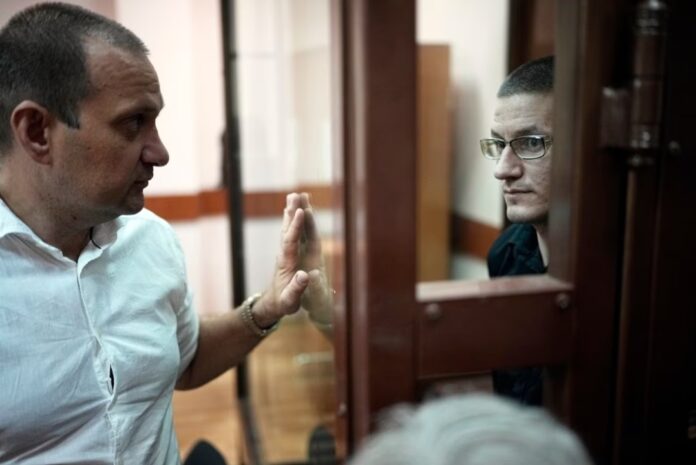 Robert Woodland, right, a Russia-born U.S. citizen, stands in a glass cage as he talks with his lawyer Stanislav Kshevitsky prior to a court hearing, Thursday, July 4, 2024, in Moscow, Russia. Woodland was convicted of drug-related charges and sentenced to 12 and a 1/2 years in prison on Thursday. (AP Photo/Alexander Zemlianichenko)